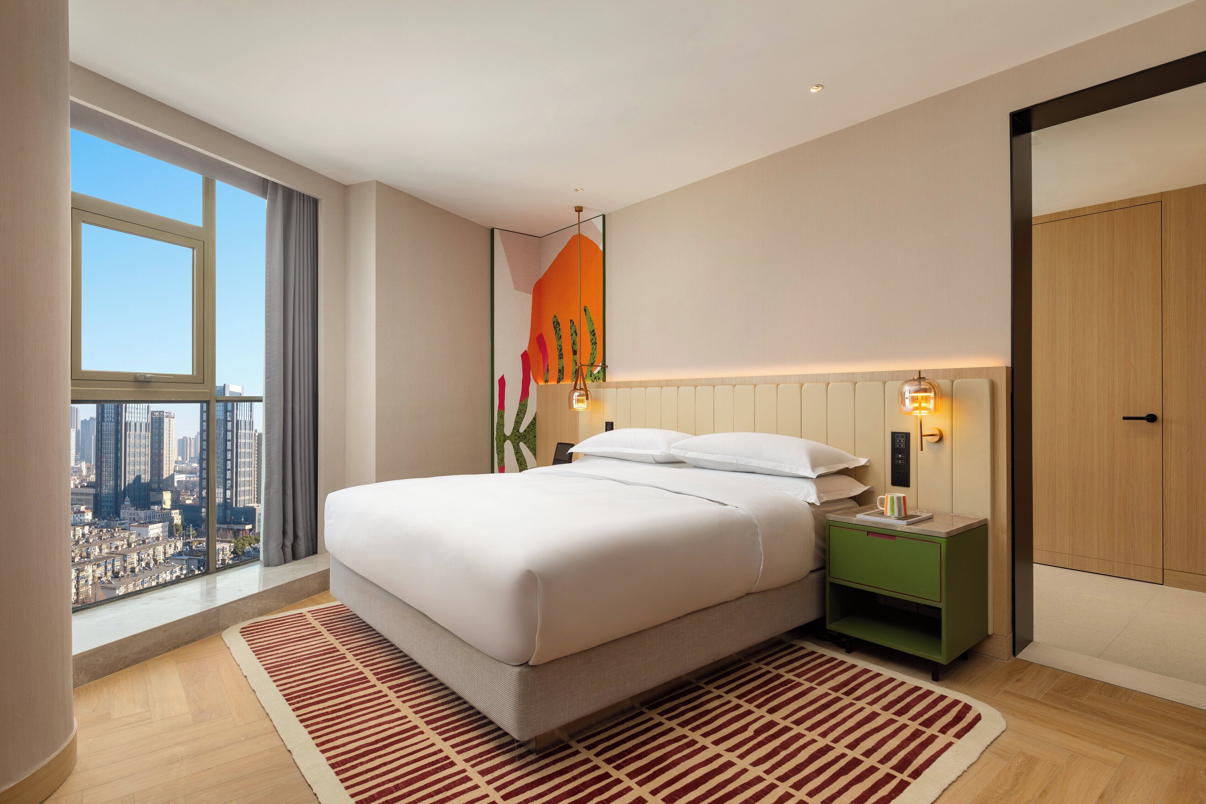 A modern hotel room with a large bed, a nightstand, a green side table, a city view, and a large window.