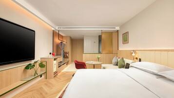 Ein modernes Hotelzimmer mit einem großen Bett, einem Flachbildfernseher, einem kleinen Schreibtisch mit Stuhl, einem runden Tisch mit Stuhl und einer kleinen Küchenzeile.