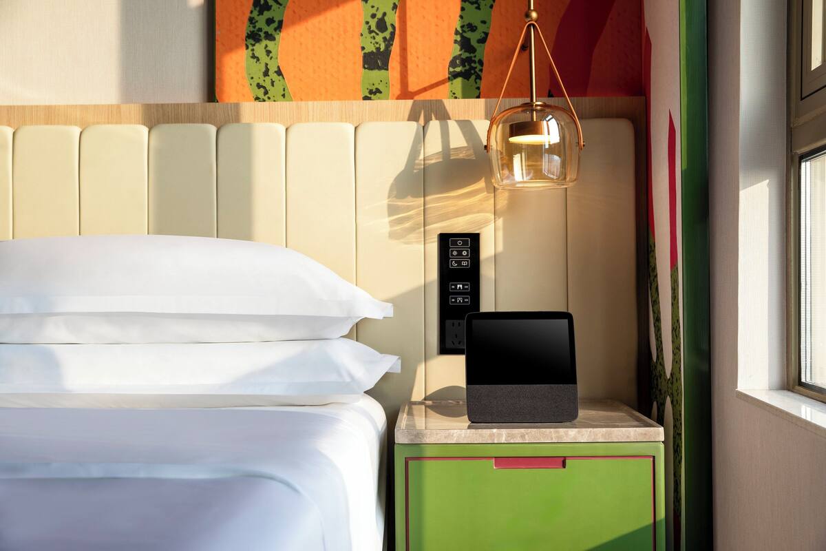 A neatly made bed with white linens, a bedside table with a tablet, a wall-mounted control panel, and a hanging light fixture.