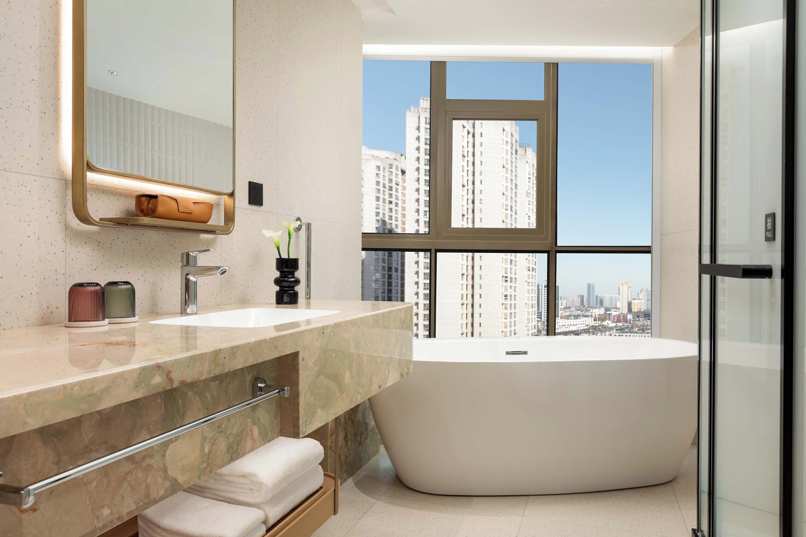 Ein modernes Badezimmer mit einer großen Badewanne, zwei Waschbecken, einem Waschtisch inklusive Spiegel und einem Fenster mit Blick auf die Stadt.