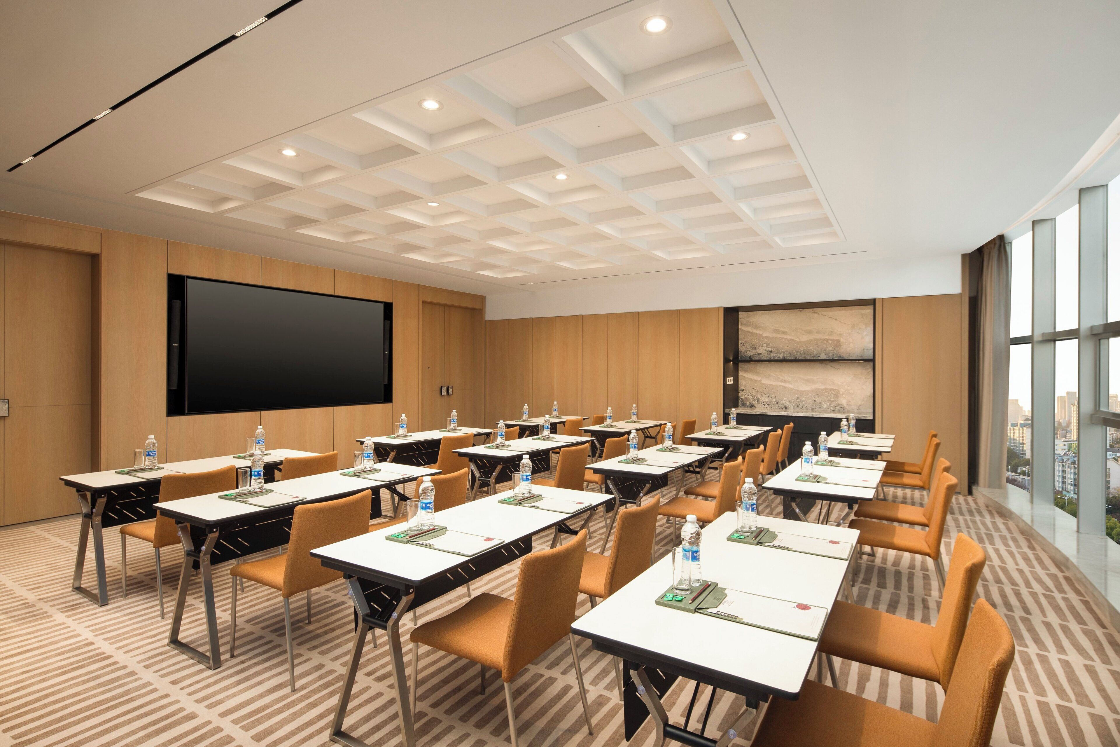 A conference room with multiple tables and chairs, a large screen, and a city view through large windows.