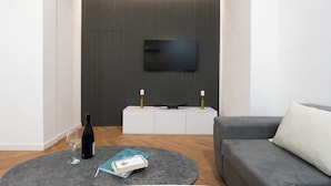 A modern living room with a flat-screen TV mounted on a textured wall, a white console table with two candles, a grey sofa, a round coffee table with a wine bottle and a book, and wooden flooring. - Chic & Modern 4BR Home in Old Tbilisi (Tbilisi)