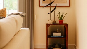 A room with a wooden floor, a beige sofa, a window with curtains, a framed beach painting, a small wooden side table with a plant, and a bowl. - Downtown Saugatuck Dream (Saugatuck)
