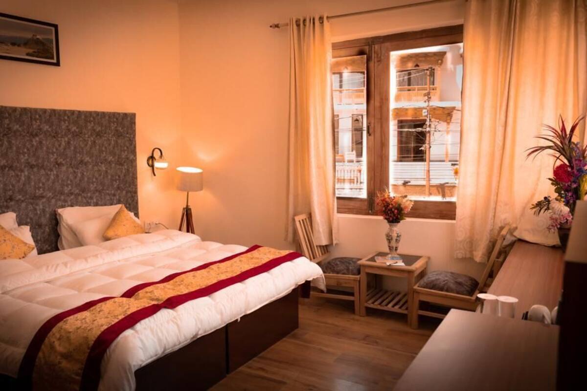 A hotel room with a large bed, a bedside table with a lamp, a chair, a small table with a vase of flowers, and a window with curtains.