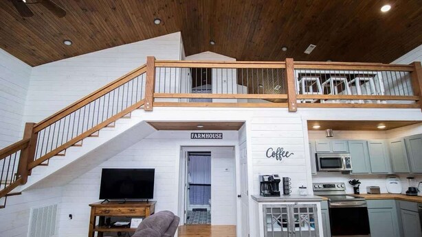 A spacious loft with a wooden staircase, a living area with a sofa and TV, a kitchen with modern appliances, and a sign indicating a farmhouse. - Ozark Escape - Hot Tub, Mountain Views, Near River (Compton)