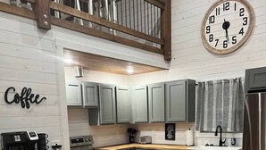A modern kitchen with grey cabinets, a wooden floor, a coffee station, and a clock on the wall. - Ozark Escape - Hot Tub, Mountain Views, Near River (Compton)