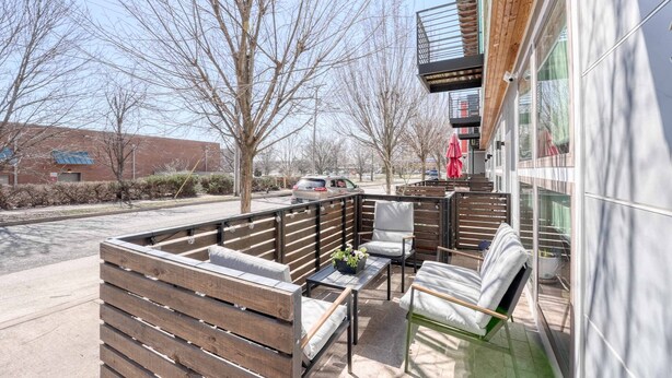 An outdoor seating area with wooden benches and chairs, a glass table, and a view of a building with large windows and a balcony. - Razorback Retreat – Walk to Stadium & Downtown (Fayetteville)
