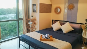 A bedroom with a large window, a bed with a dark blue bedspread, and a wooden nightstand with a lamp. - BGC McKinley Center Prime Accommodations (Taguig)