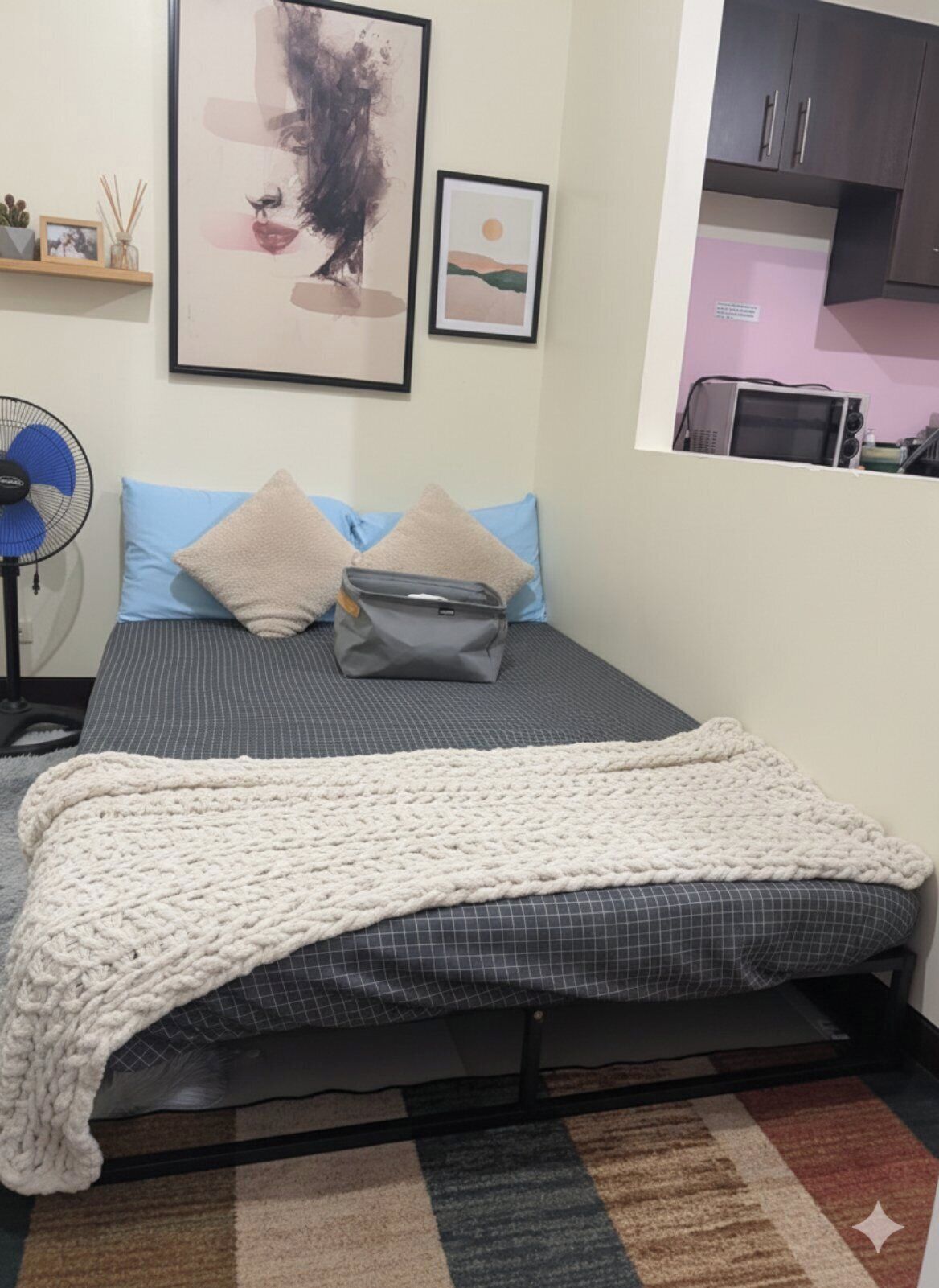 Une chambre à coucher de taille réduite, comprenant un lit, des oreillers, une couverture, un ventilateur et un petit coin cuisine.