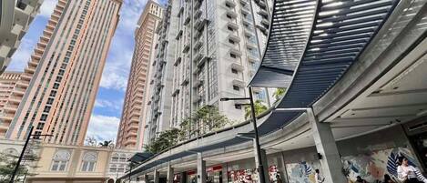 A modern urban street with tall residential buildings, a covered pedestrian walkway, and a commercial area with shops.