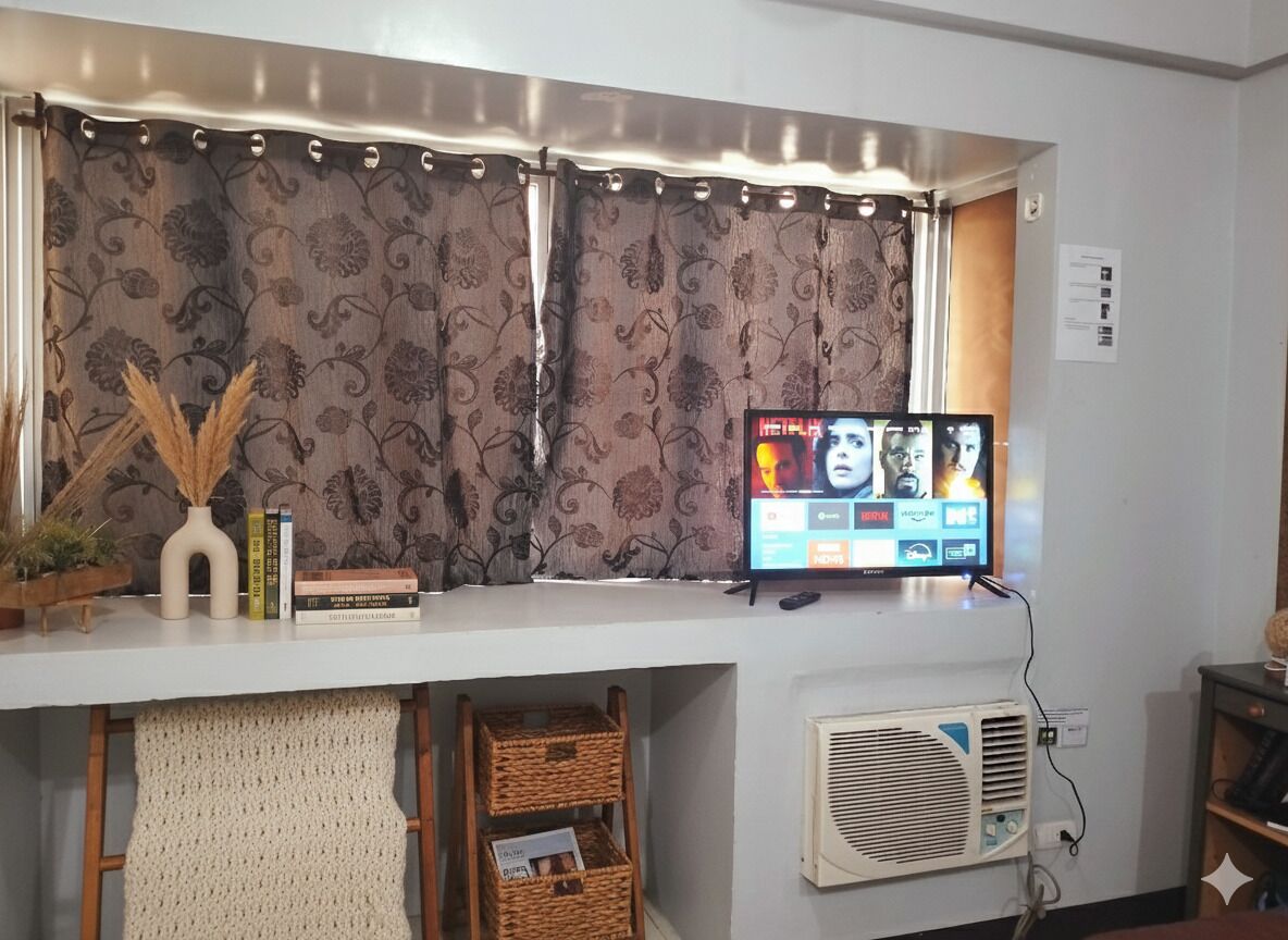 A room with a flat-screen TV mounted on the wall, a window with curtains, a small table with books and decorative items, and a wall-mounted air conditioning unit.