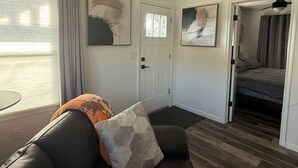 Un salon moderne avec un canapé en cuir noir, une table basse en verre et des œuvres d’art abstraites accrochées au mur. Une porte donne accès à une chambre à coucher équipée d’un lit et d’un ventilateur de plafond.