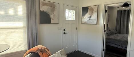 Una sala moderna con un sofá de cuero negro, una mesa de centro de vidrio y arte abstracto en la pared. Una puerta conduce a un dormitorio con cama y ventilador de techo.