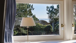 Una ventana con una lámpara sobre una superficie blanca, una cortina y vistas a árboles y un cuerpo de agua.