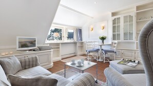 Una sala de estar acogedora con sofá, sillones, una mesa de centro y televisor. Hay una zona de comedor con mesa y sillas, y una cocina con armarios y ventana.