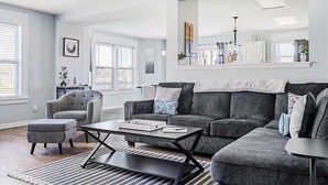 A modern living room with a grey sofa, a matching armchair, and a coffee table. There is a large mirror above the sofa and a bookshelf with various items. - The Cozy Charcoal Cottage | Garage | Grill & Patio (Brownsburg)