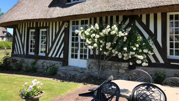 A traditional half-timbered house with a thatched roof, white window frames, and a garden with white flowers and a metal chair.