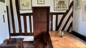 A room with exposed wooden beams, a wooden door, a tiled floor, a wooden table with a bowl, and framed pictures on the walls.