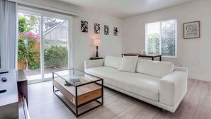 A modern living room with a white sofa, a glass coffee table, and a TV stand. There are abstract wall art pieces and a lamp on a side table. - Oceanview Condo 3 Blocks to La Jolla Beach (San Diego)