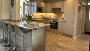 A modern kitchen with a central island, bar stools, a microwave, oven, and a large window. - Stylish, Contemporary Home in Earl Shilton (Leicester)