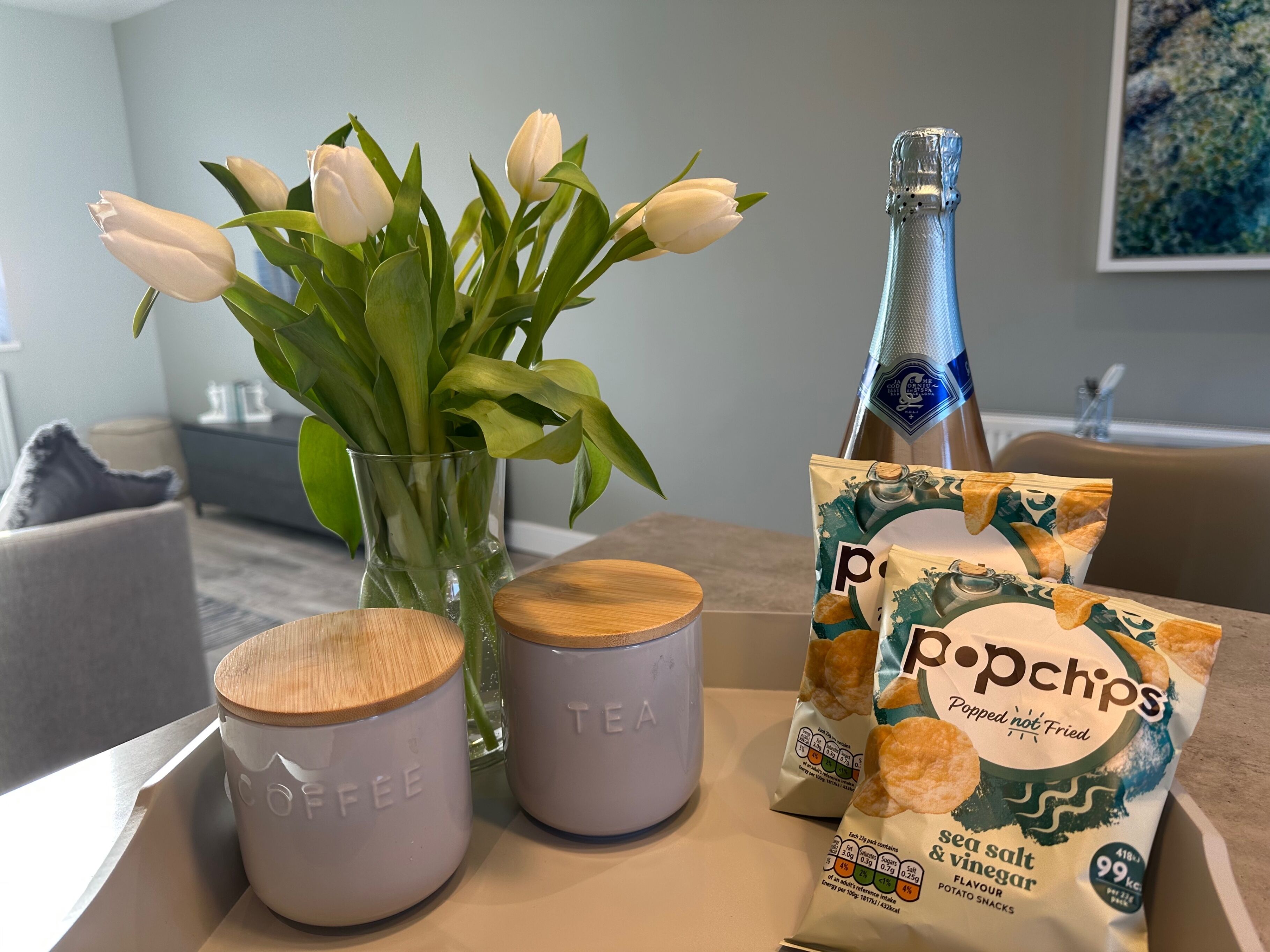 Une table avec un vase de tulipes blanches, deux tasses à café, une bouteille de champagne et un paquet de chips.