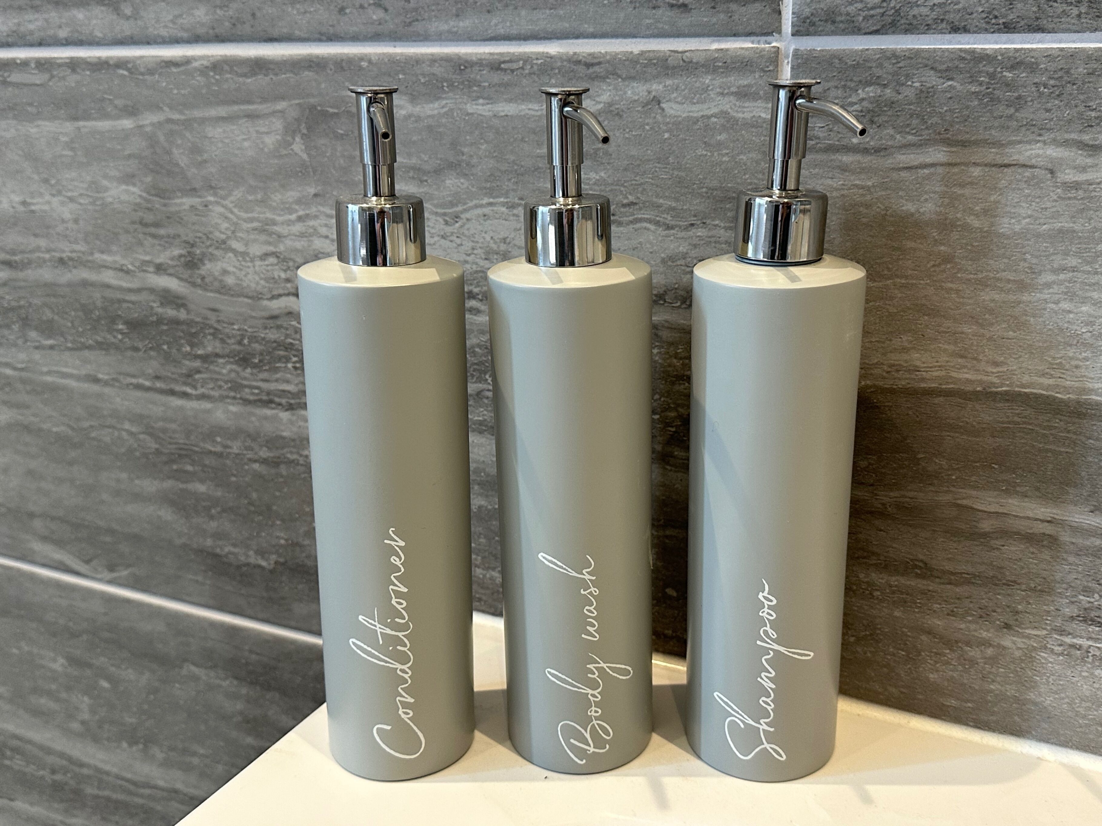 Three cylindrical bottles with pumps, labeled 'Conditionner,' 'Body wash,' and 'Shampoo,' placed on a white surface against a grey tiled wall.