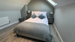 A bedroom with a sloped ceiling, a skylight, a bed with a grey blanket, two bedside tables with lamps, and a radiator. - Stylish, Contemporary Home in Earl Shilton (Leicester)