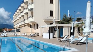 Una piscina con sillones de descanso y sombrillas, un edificio con balcones y un cielo despejado.