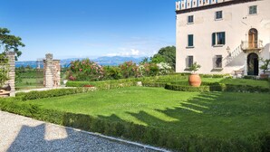 A historic building with a well-maintained garden, a stone gate, and a view of the sea and mountains in the background.