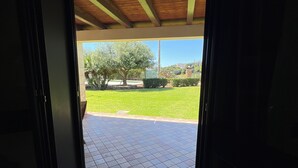 Une porte-fenêtre donnant sur un patio extérieur, doté d’un sol carrelé et offrant une vue sur un arbre et une pelouse.