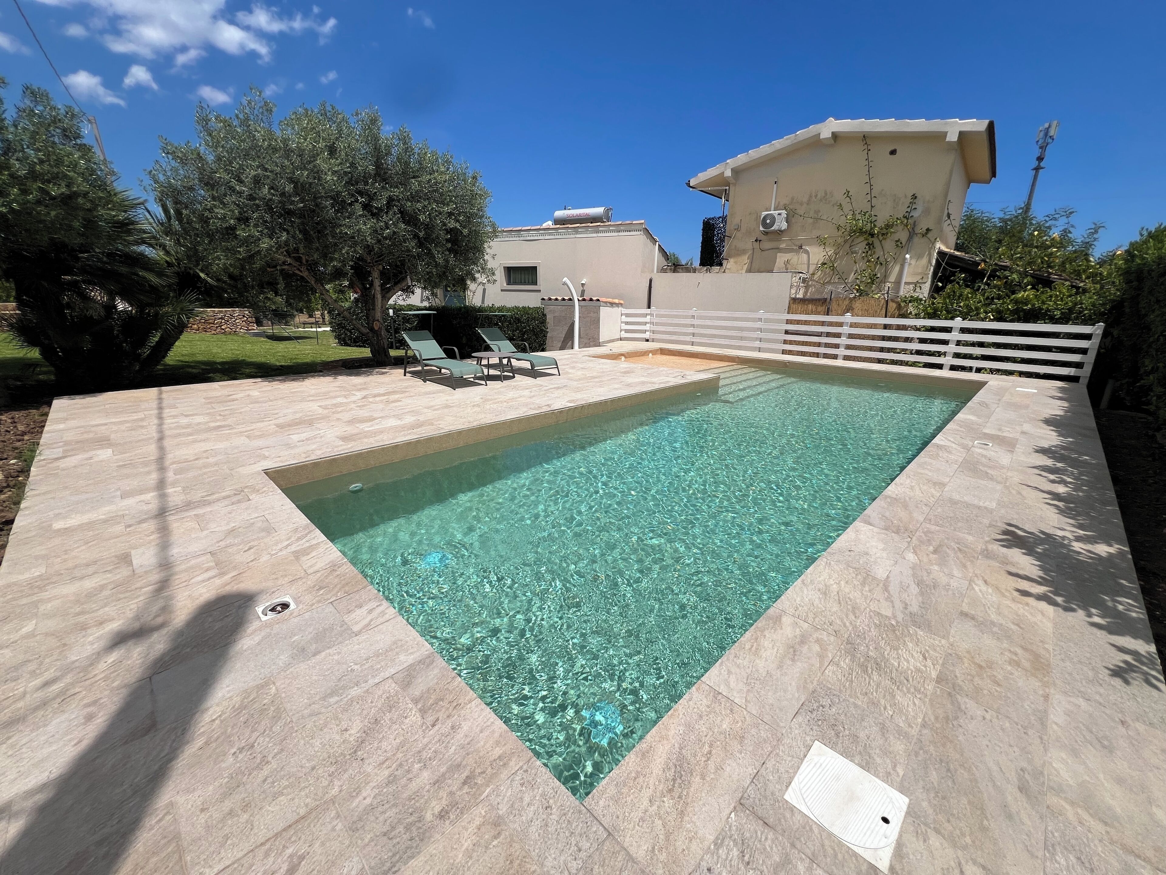 A rectangular swimming pool with clear water, surrounded by a tiled deck. There are two lounge chairs and a small table on the deck.