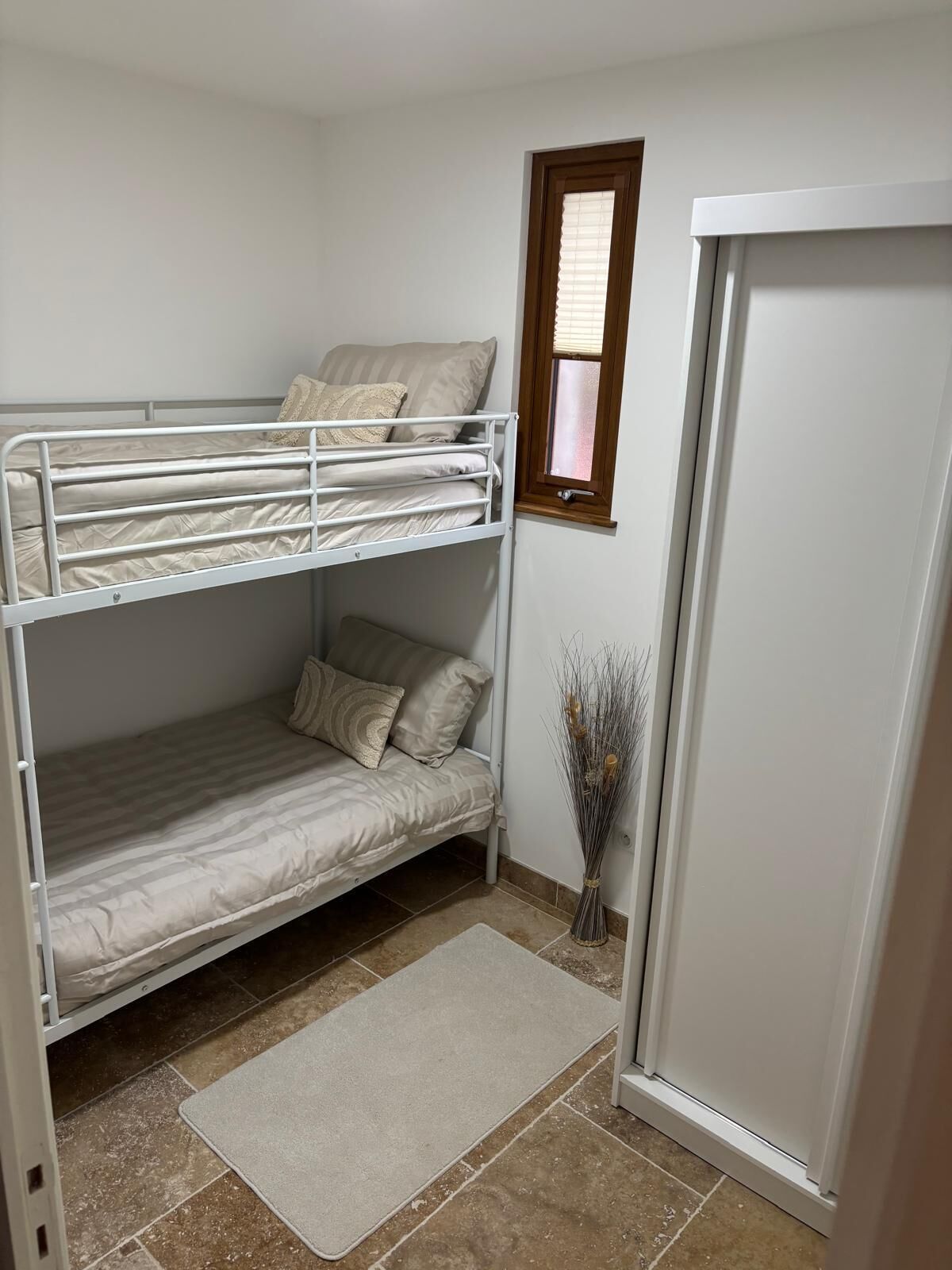 Une chambre avec un lit superposé, une fenêtre, une porte et un vase décoré de branches.