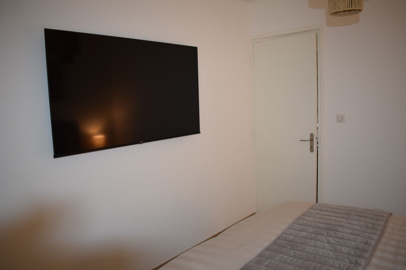 A bedroom with a bed, a wall-mounted TV, a door, and a light fixture.