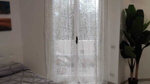 Ein Schlafzimmer mit einem Bett, einem großen Fenster mit Vorhang, einer Zimmerpflanze und einer Klimaanlage.