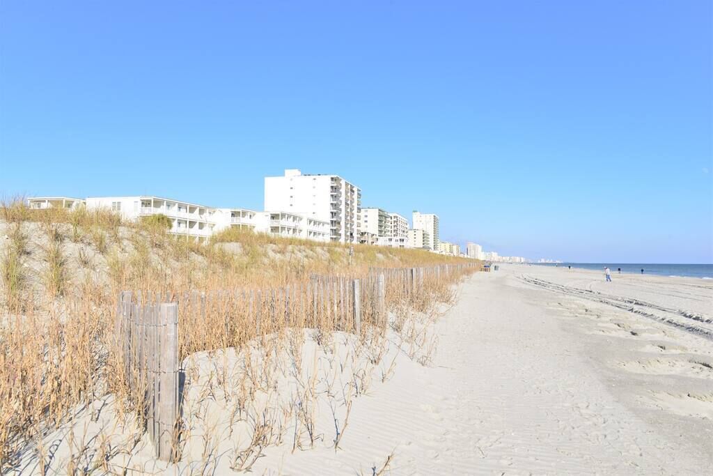 A sandy beach with a wooden fence and buildings in the background.