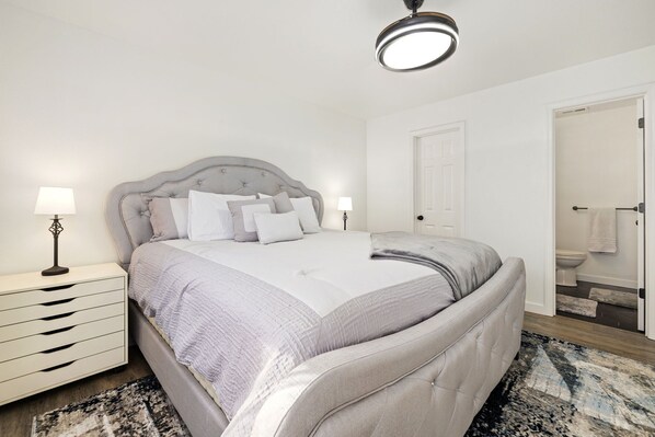 Un dormitorio con una cama grande, mesitas de noche, una lámpara de techo y una puerta que da a un baño.