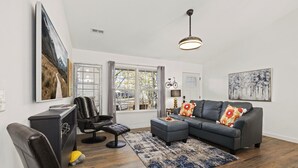 Una sala de estar con sofá, reposapiés y sillón. Hay una ventana con cortinas, una lámpara y obras de arte en las paredes.