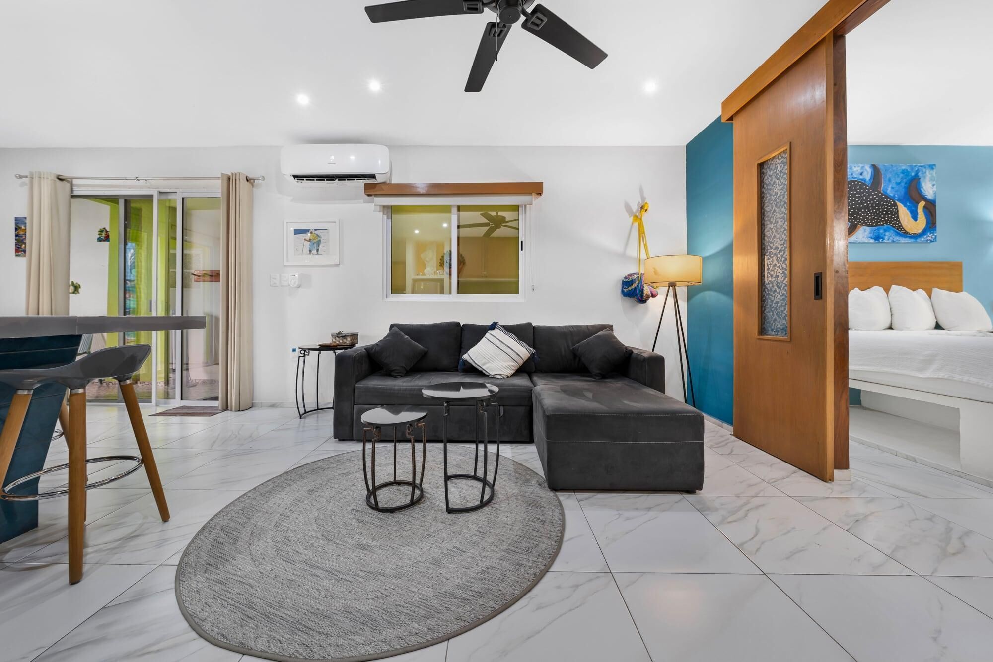 Una sala moderna con un sofá seccional, un tapete redondo, un comedor con mesa y sillas, y un dormitorio con cama y un cuadro.