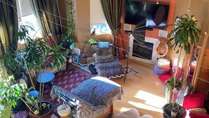 A cozy living room with a wooden sofa, a patterned ottoman, and a fluffy rug. There are several potted plants, a television, and a painting on the wall. - Romantic Plant filled studio Beautiful vibes! Spa Services Available (Emeryville)