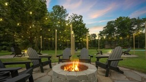 A stone fire pit surrounded by Adirondack chairs in a landscaped outdoor area with trees and string lights.