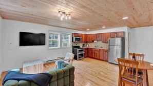 A spacious living area with a wooden ceiling, a flat-screen TV, a sofa, a dining table with chairs, and a kitchen with wooden cabinets and appliances. - Charming Pocono Boutique | Firepits | Pickleball (Monroe County)