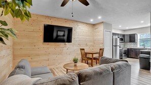 A modern living room with a wooden wall, a flat-screen TV, a gray sofa, a small round table with a plant, and a ceiling fan.