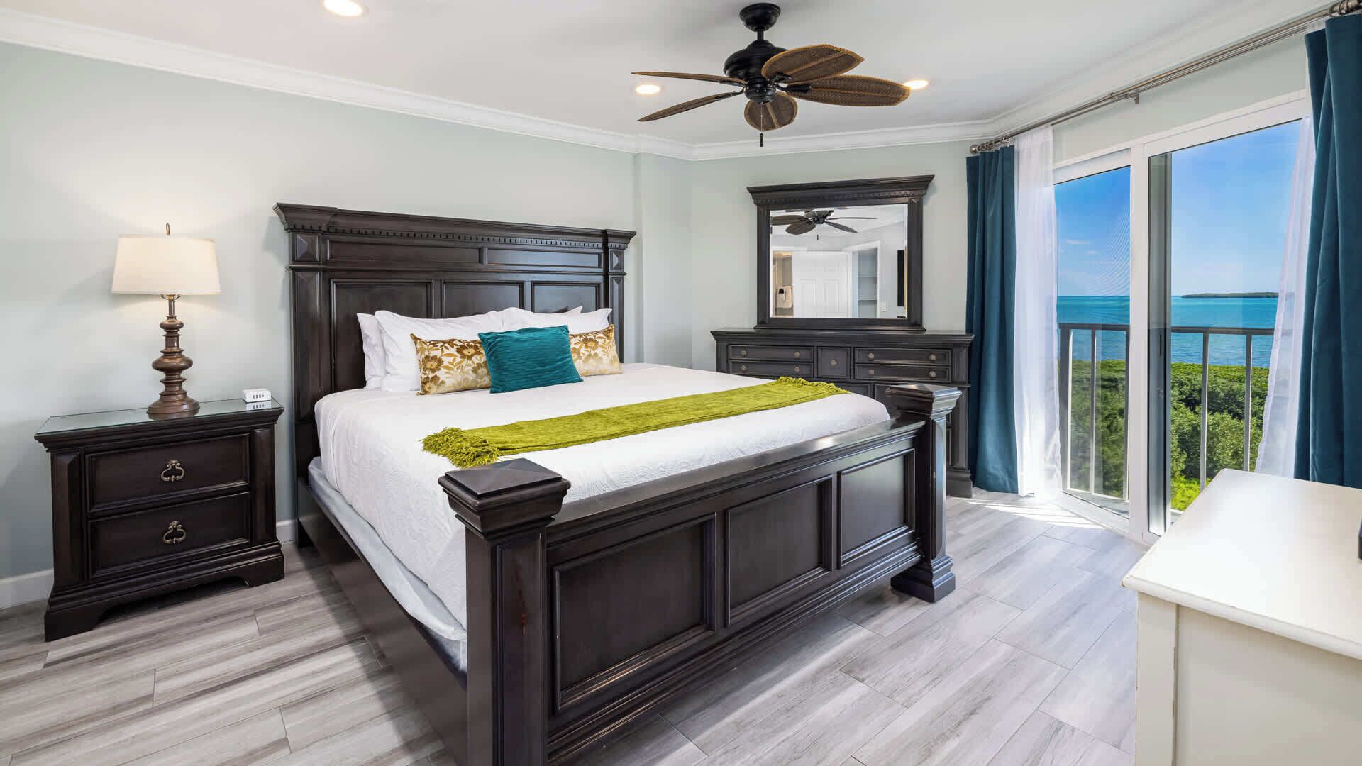 A bedroom with a large bed, dark wood headboard, a nightstand with a lamp, a dresser, a large mirror, and a balcony with a view of the ocean.