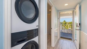 Un cuarto de lavado con lavadora y secadora de carga frontal, una puerta que da a un balcón con vistas a palmeras y cielo despejado.