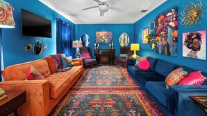 A vibrant living room with a blue wall, a ceiling fan, a flat-screen TV, a fireplace, and a colorful rug. - Boho Bougie: Pool, Hot Tub & Epic Back Yard (Fredericksburg)