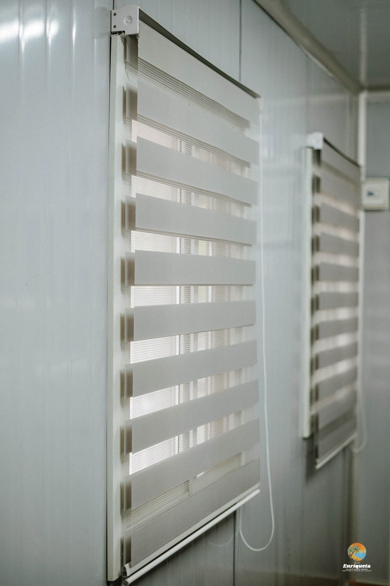 A room with two white blinds on a wall, a visible electrical outlet, and a door handle.