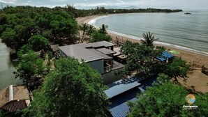 Una propiedad costera con vista a la playa, una casa grande con techo metálico y un pequeño bote en el agua.