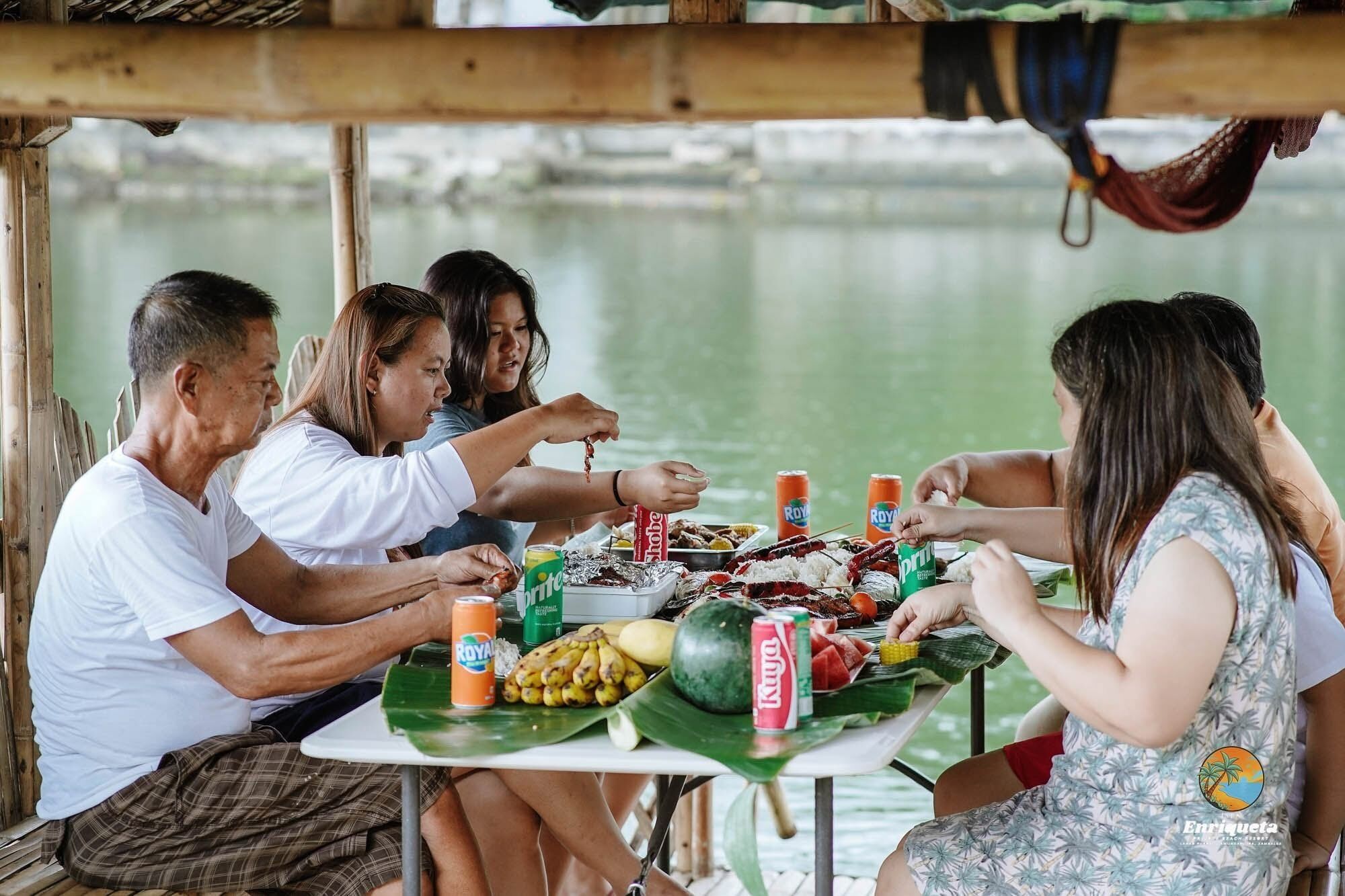 A group of people dining at a table with a view of a body of water, featuring a variety of food items and drinks.