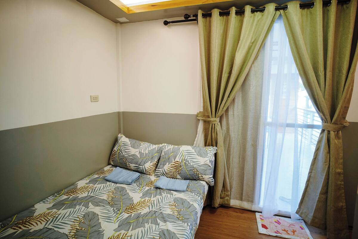 A small, neatly made bed with a leaf-patterned bedspread and pillows, a window with sheer curtains, and a wooden floor.