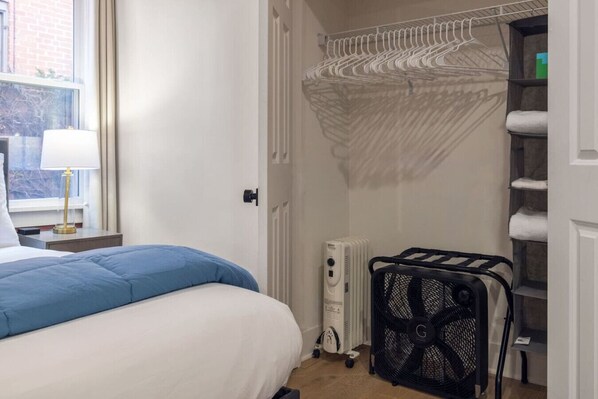 A hotel room with a bed, a radiator, a laundry cart, and a window with a view of a brick building.
